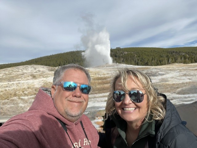 Cindy and Lee at Yellowstone with a geyser erupting behind them